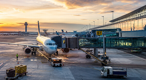 Airplane parked at an airport terminal gate B1 with a boarding bridge attached, ground crew and luggage carts nearby, and a control tower in the sunset sky