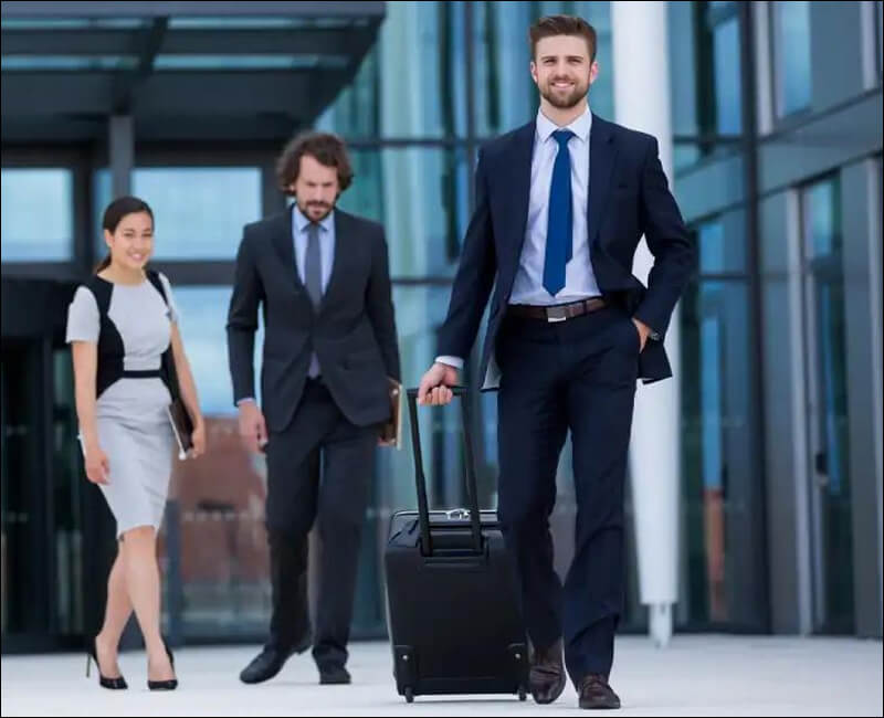 Business professionals in suits walking outside a modern glass office building, with the man in front pulling a rolling suitcase