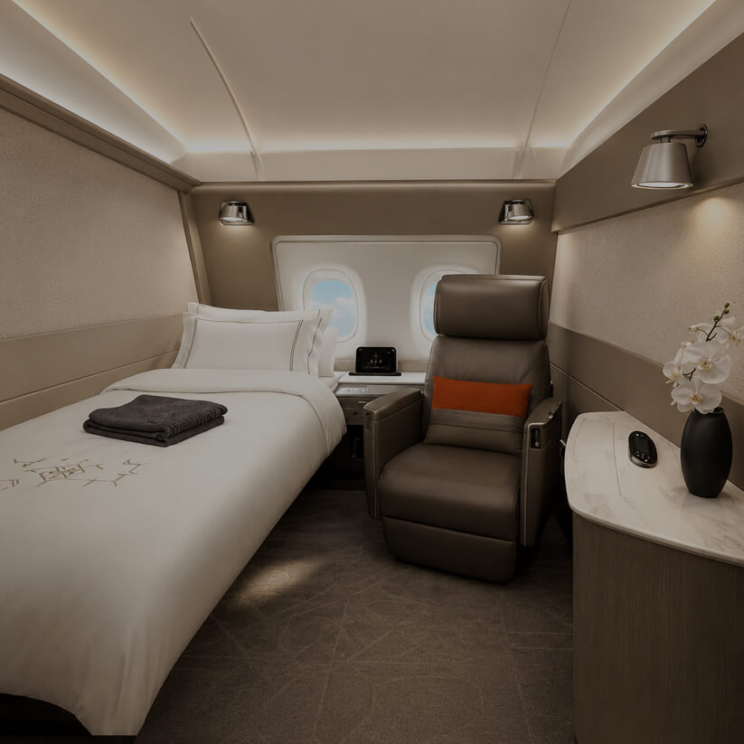 First class airline suite with a made-up bed, brown leather lounge chair with orange cushion, side table, and a marble console with orchid flowers