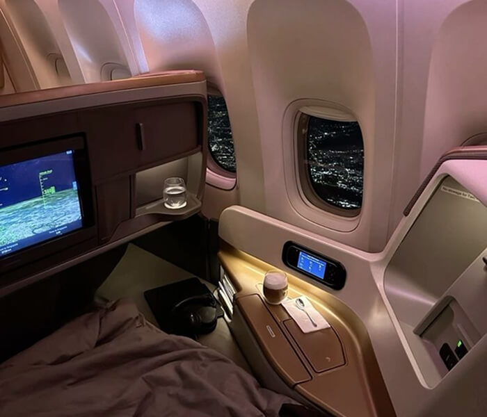 First class airline suite at night with a lie-flat bed, entertainment screen showing the flight map, and city lights visible through the window