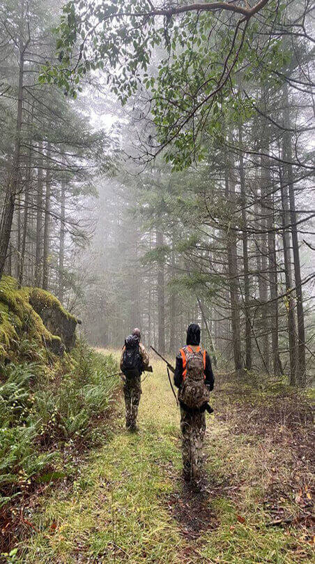 Two hunters in camouflage with backpacks and rifles walking down a foggy forest trail lined with tall trees