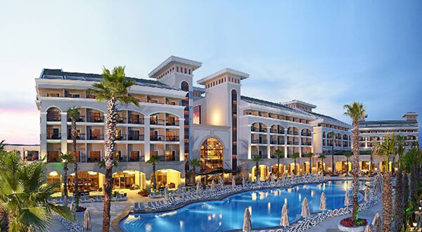 Luxury resort hotel exterior lit up at dusk with palm trees, sun loungers, and a large outdoor pool in front of the building