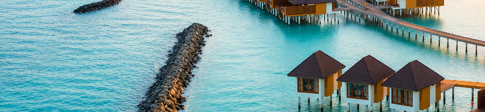 Aerial view of overwater bungalows connected by wooden walkways above a turquoise lagoon with a rock breakwater nearby