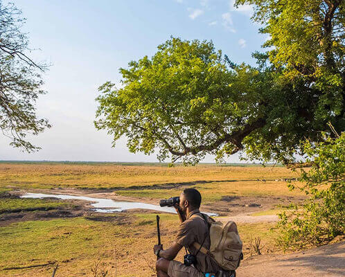 Man with a backpack and camera on a tripod overlooking an open African savanna with a waterhole and trees