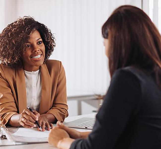 Smiling businesswoman in a blazer discussing documents with another woman across a desk with a laptop during an office meeting